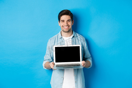帅气的年轻人在笔记本电脑屏幕上介绍产品，展示电脑，微笑，站在蓝色背景上