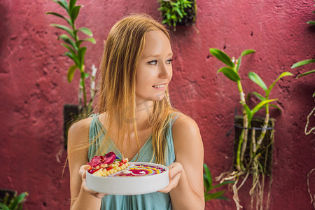 年轻女人吃地中海早餐，吃健康的热带早餐，冰沙碗和热带水果，装饰着彩色酸奶、姜黄和螺旋藻的图案。