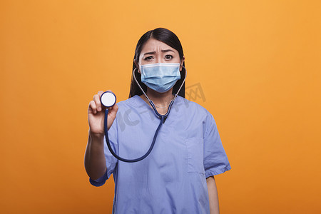 护士戴口罩摄影照片_担心的医疗诊所亚洲护士拿着听诊器戴着防护面罩。