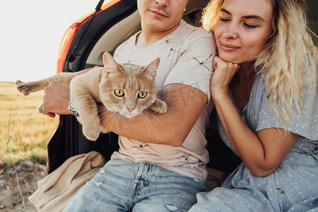 年轻男女和宠物坐在汽车后备箱里，幸福的夫妻和他们的红猫享受公路旅行