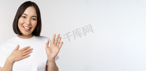 美丽的韩国女孩举手的画像，自我介绍，手放在心上，问候，站在白色背景上