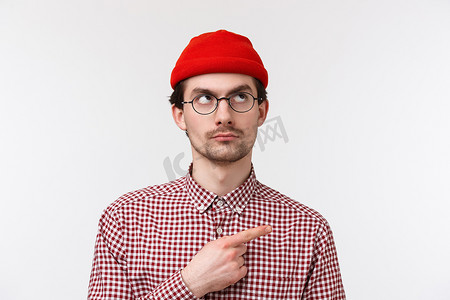 帽子创意摄影照片_身穿红色帽子和格子衬衫、眼镜、怀疑地偷看、表情严肃地指着右上角、站在白色背景上的持怀疑态度的帅气年轻嬉皮士