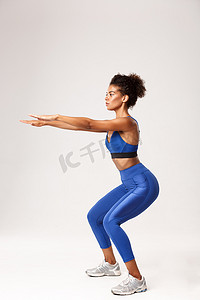 坚定的非洲裔美国健身女性的全长侧面照片，身穿蓝色运动服，双手向前伸直蹲下，在白色背景下锻炼