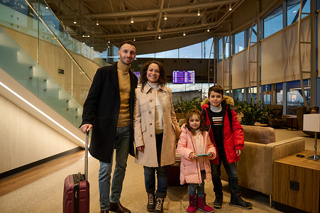 高兴快乐的孩子和享受家庭旅行的父母，带着手提箱站在机场出发航站楼的休息区，对着航班信息面板和时间表。