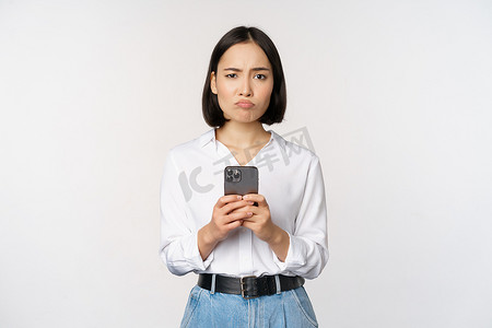 亚洲女性手持智能手机，疑惑地看着，对手机应用程序感到失望，站在反对的白色背景下