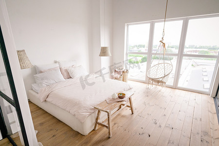 宽敞明亮的卧室采用柔和的奶油色简约风格，配有一张双人床、白色墙壁、柳条洗衣篮、柳条枝形吊灯、米色床长椅、大窗户、波西米亚风吊椅和木地板。