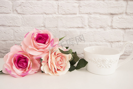 粉色漱口杯摄影照片_白色桌子上的玫瑰花束，前面天使的一大杯咖啡，浪漫的花框背景，花卉风格的墙壁模型，玫瑰花模型，情人节母亲节卡，礼品卡