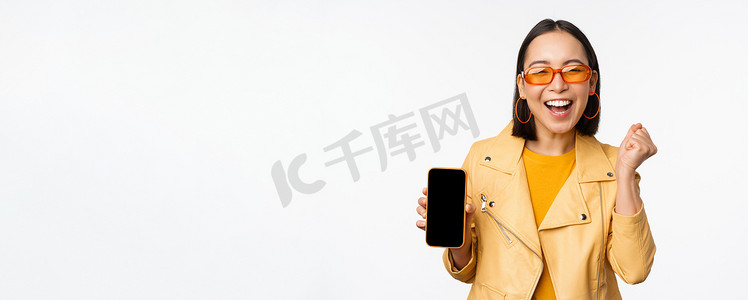 app展示界面摄影照片_戴墨镜的快乐亚洲女孩，展示手机屏幕、智能手机界面，笑着笑着，庆祝着，站在白色背景上