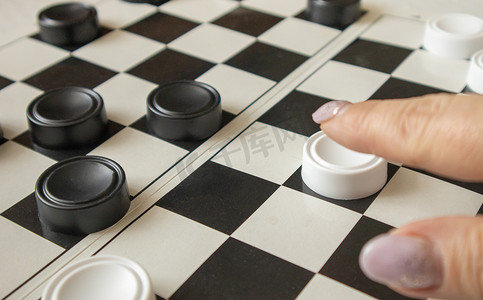 跳棋棋子摄影照片_一个女人的手在黑白运动场上移动一个白色棋子，这是爱好和家庭游戏的概念