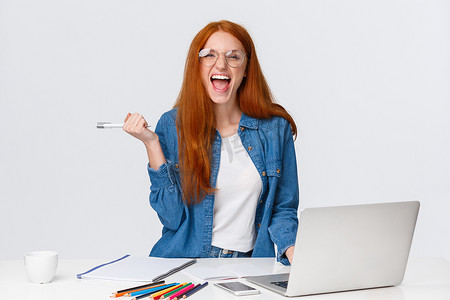 兴奋而胜利的漂亮红发女学生通过了挑战，使用笔记本电脑在线发现期末考试评分，拿着笔，快乐地尖叫，站在白色背景