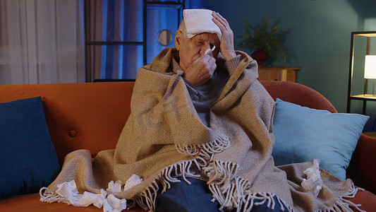 老人流感摄影照片_患有感冒或过敏症的老祖父，把鼻涕吹到坐在家里的餐巾纸上