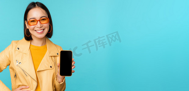 戴着时尚太阳镜的年轻时尚亚洲女模特的形象，显示手机屏幕，智能手机界面上的应用程序，站在蓝色背景上