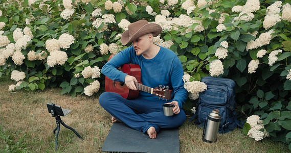 带着牛仔帽摄影照片_带着吉他的男旅行者，坐在大自然的公园里，进行在线广播