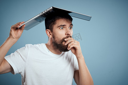 公众号台头摄影照片_情绪化的男人，头顶上有一台打开的笔记本电脑 在蓝色背景中，穿着白色 T 恤，裁剪视图