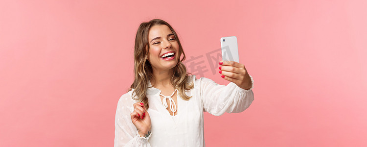开朗乐观的金发女孩特写肖像，穿着白色连衣裙，在录制视频时大笑，在移动应用程序上给朋友打电话，拍照，在智能手机上自拍，粉红色背景
