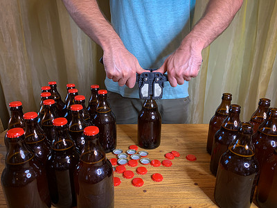 啤酒瓶啤酒盖摄影照片_男人在家酿造精酿啤酒，用塑料封盖器在木桌上用红色皇冠盖封闭棕色玻璃啤酒瓶。