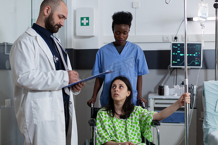 静脉注射摄影照片_护士坐在轮椅上，拿着静脉注射杆，而医生则拿着剪贴板，为患有疾病的妇女提供护理