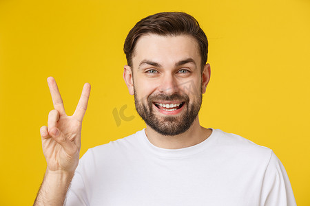 黄色手指手势摄影照片_年轻英俊的男子穿着条纹 T 恤，在孤立的黄色背景上微笑着看着镜头，手指在做胜利的手势。