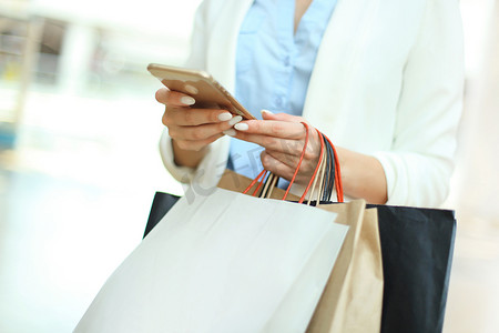 商场营销摄影照片_站在商场背景上使用智能手机和提着购物袋的女人。