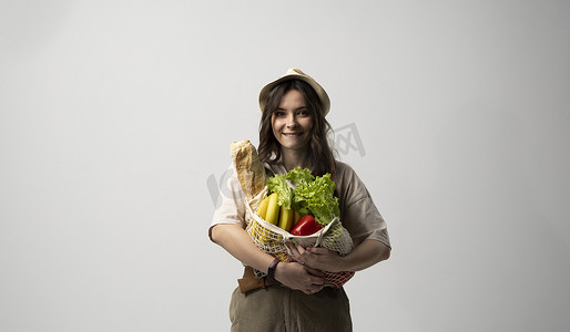 穿着米色超大号 T 恤的环保积极的年轻女性拿着可重复使用的网状棉质生态袋，在白色背景下与有机食品一起购物。