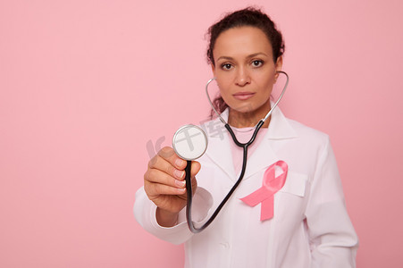 国际卫生组织摄影照片_专注于身穿粉红色缎带医用大衣的美丽混血西班牙裔医生手中的语音内窥镜，这是 10 月 1 日全球乳腺癌宣传日的象征。