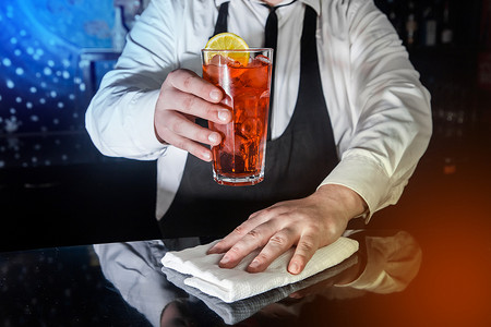 一位专业的酒保手里拿着一杯红色的酒精鸡尾酒，一杯冰镇饮料，上面装饰着一片柠檬片，并用白布擦拭吧台，供上菜