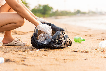 黑色垃圾塑料袋摄影照片_为清洁海滩而将塑料瓶捡进黑色垃圾塑料袋的志愿者妇女