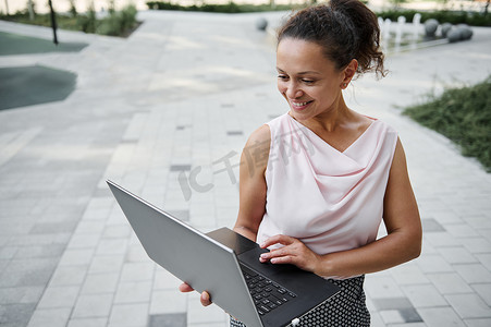 华丽的西班牙裔年轻商业自由职业者女性在笔记本电脑上工作时可爱地微笑着露齿微笑，站在城市建筑背景上。