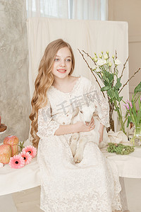女孩坐在复活节餐桌旁，手里拿着蛋糕、春花和鹌鹑蛋，怀里抱着一个白人山羊。