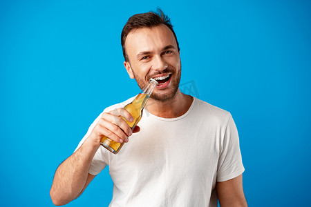 啤酒瓶啤酒盖摄影照片_蓝色背景下，身穿 T 恤、拿着啤酒瓶的快乐年轻人