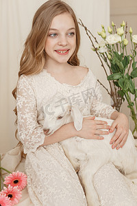 抱着花的女孩摄影照片_女孩坐在复活节餐桌旁，手里拿着蛋糕、春花和鹌鹑蛋，怀里抱着一个白人山羊。