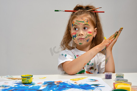 穿着白色 T 恤、头发上梳着刷子的小女孩坐在桌边，手里拿着什么东西和油漆，在上面画画。