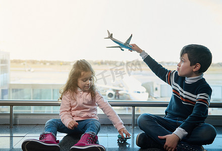 可爱的孩子、男孩和小女孩坐在地板上玩玩具飞机，全景窗户俯瞰国际机场出发航站楼的跑道和飞机