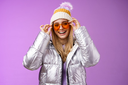 下雪保暖摄影照片_漂亮迷人、快乐、微笑的金发女友在度假时玩得很开心，女朋友戴上太阳镜，咧着嘴笑，高兴地穿着凉爽的银色闪闪发光的夹克，保暖的冬帽，紫色背景