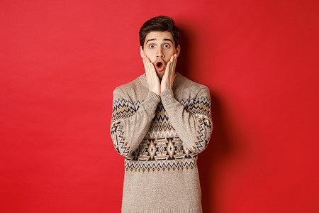 惊讶的帅哥对酷炫的新年促销活动做出反应，惊讶地喘着粗气，穿着圣诞毛衣，站在红色背景上