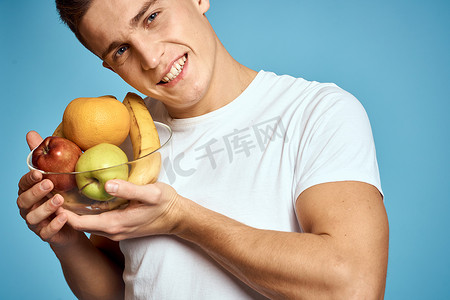 快乐的男人，手里拿着新鲜水果，双手蓝色背景白色T恤维生素香蕉橙子苹果