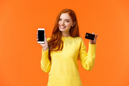 红发白人女孩在智能手机上展示应用程序，如手持手机和信用卡，使用移动银行应用程序，笑得开心的相机，在橙色背景下做广告