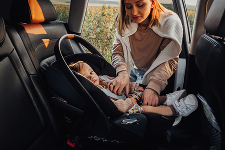 驾驶的摄影照片_年轻的母亲把她的小女儿放在车里的儿童汽车座椅上