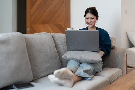 身穿蓝色运动衫的快乐年轻女性在家里工作，坐在蓝色沙发上，沙发上配有笔记本电脑和猫，远程工作和教育，待在家里