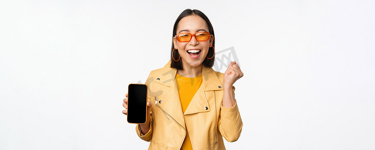 手机界面摄影照片_戴着太阳镜的快乐亚洲女孩，展示手机屏幕、智能手机界面，笑着，庆祝，站在白色背景上