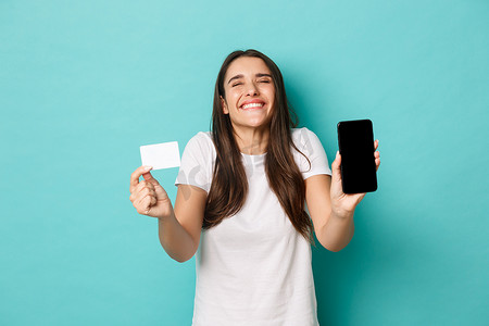 身穿白色 T 恤的幸运女性肖像，心情愉快，微笑着，用手机出示信用卡，站在蓝色背景上