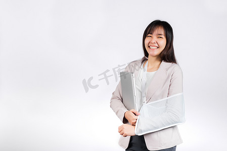 亚洲女商人在事故后自信地微笑着断臂，并戴上手臂夹板进行治疗，但仍然必须工作，她拿着笔记本电脑