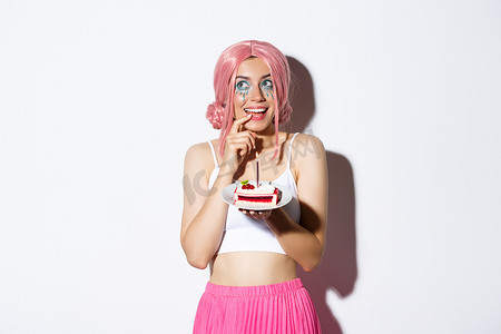 戴着粉色派对假发的可爱生日女孩的画像，拿着蛋糕，在许愿前思考，站在白色背景上