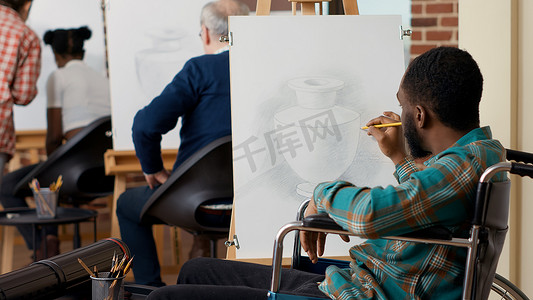 轮椅设计摄影照片_坐在轮椅上的艺术班学生用铅笔在画布上画花瓶