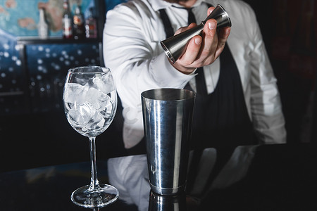 专业酒保的手将糖浆倒入金属工具中的量杯中，用于准备和搅拌振动器饮料的酒精鸡尾酒，以及带冰块的玻璃杯