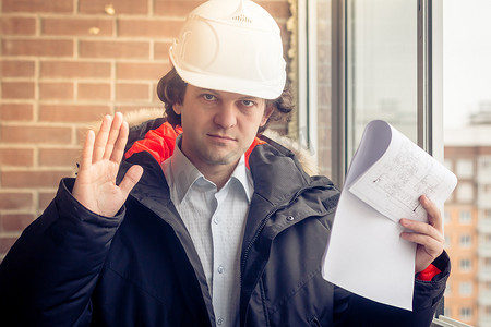 一名愤怒不满的建筑工人戴着头盔，一手拿着项目图纸，另一只手拿着手机，对着某人尖叫。