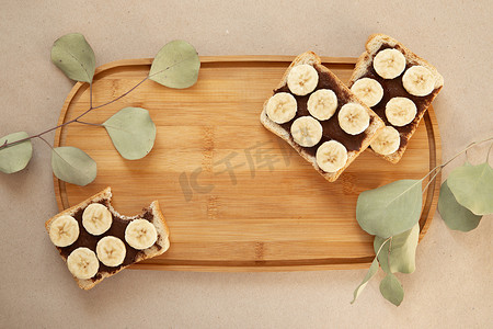 三块涂有巧克力黄油的香蕉白面包吐司放在一块切菜板上，牛皮纸背景上有一枝叶子。