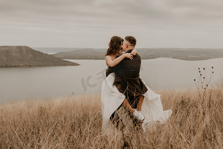恩爱的情侣婚礼新婚夫妇在礼服面纱运动鞋和西装拥抱亲吻在高高的草地上在河流上方的山上旋转