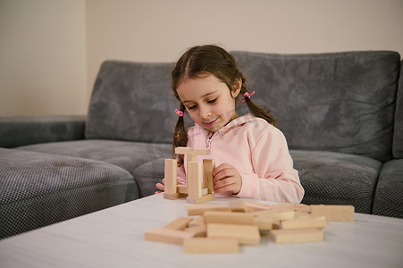 欢快、有创意的漂亮白人女婴，有两条辫子，穿着粉色运动衫，玩教育发展棋盘游戏，她专注于用积木建造木结构