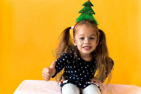 正面微笑的肖像惊讶快乐的小女学生女孩圣诞树装饰在头上的圆点连衣裙上竖起大拇指橙色背景。